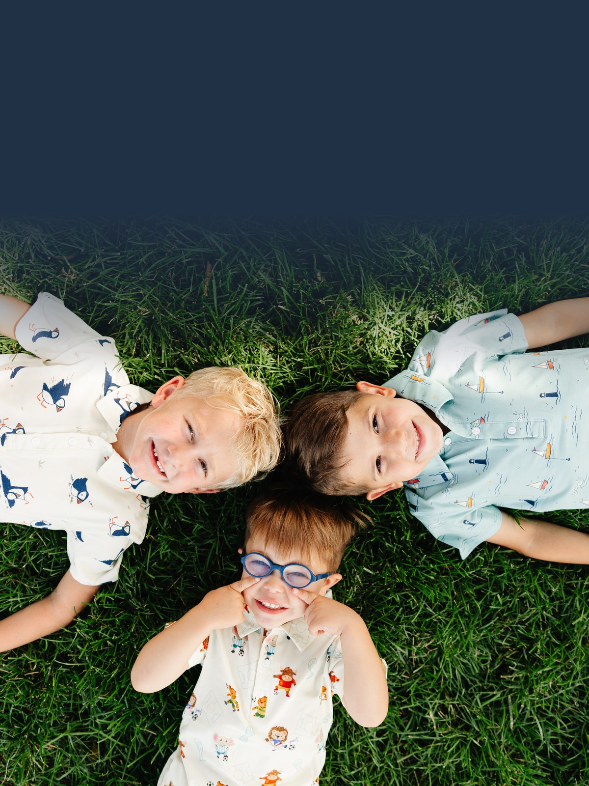 Three young boys lying in the grass smiling up at the camera, wearing Hugs & Giggles puffin, sailboat, and Zoo Champions soccer print shirts.