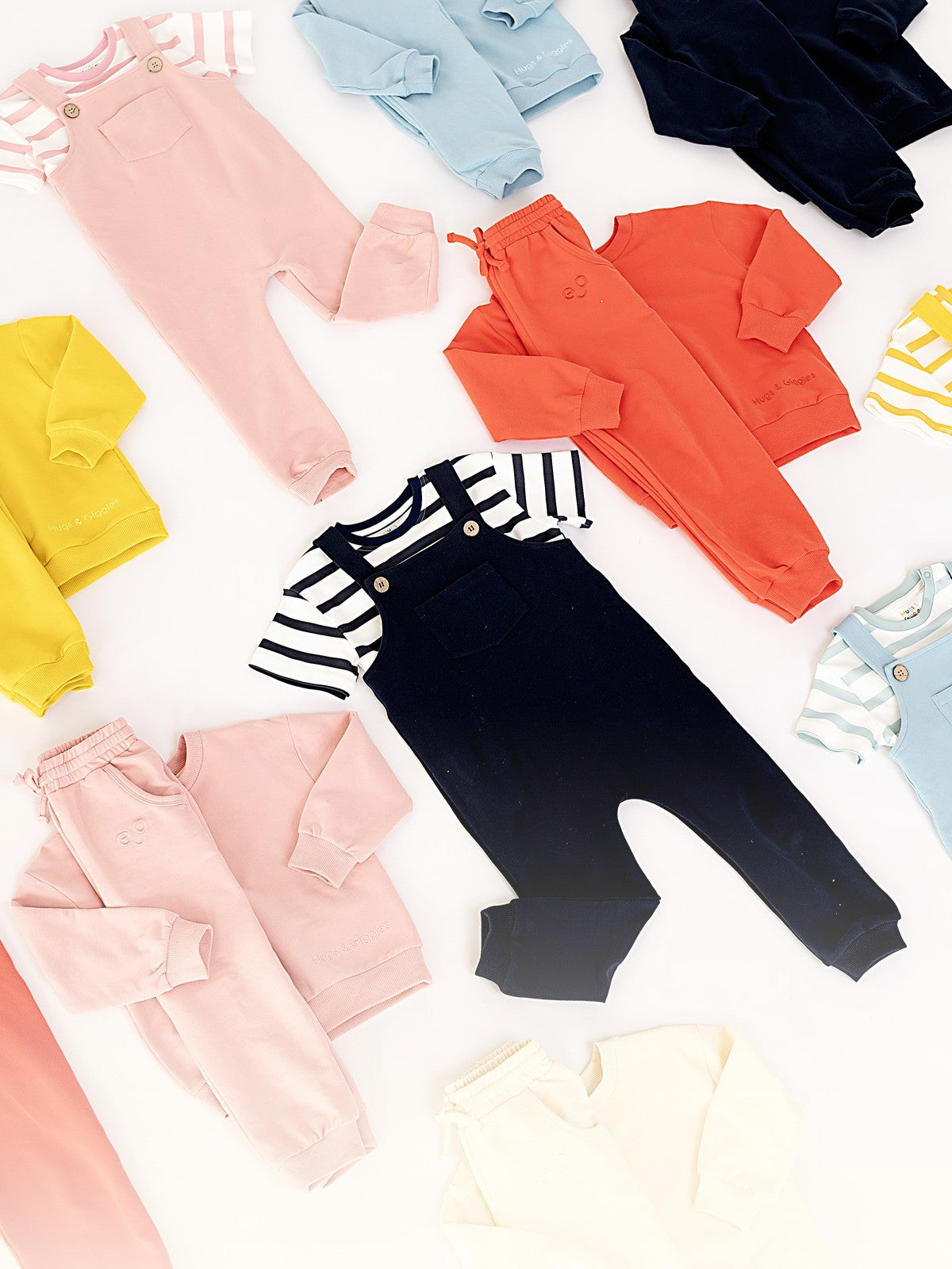 Flat lay of Hugs & Giggles French Terry organic cotton overalls and sweatshirt sets in yellow, pink, coral, navy, blue, and cream.