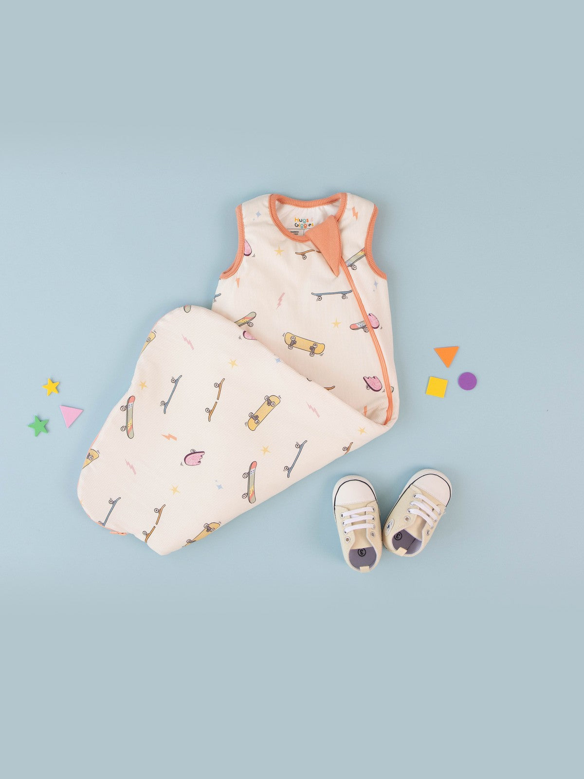 Hugs & Giggles skateboard print sleep bag laid flat on a soft blue backdrop with baby sneakers and colourful shapes.