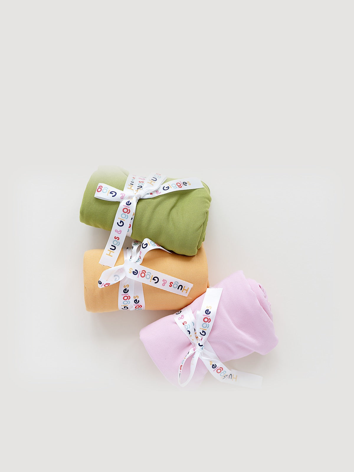 Three rolled Hugs & Giggles ribbed swaddle blankets in olive green, peach, and soft pink, each tied with a branded Hugs & Giggles ribbon.